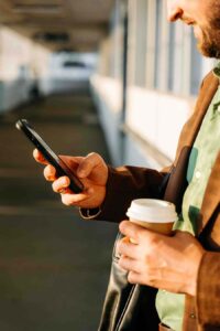 A men holding and looking to his phone and holding a cup of coffee to go somewhere outside - Carrier Billing